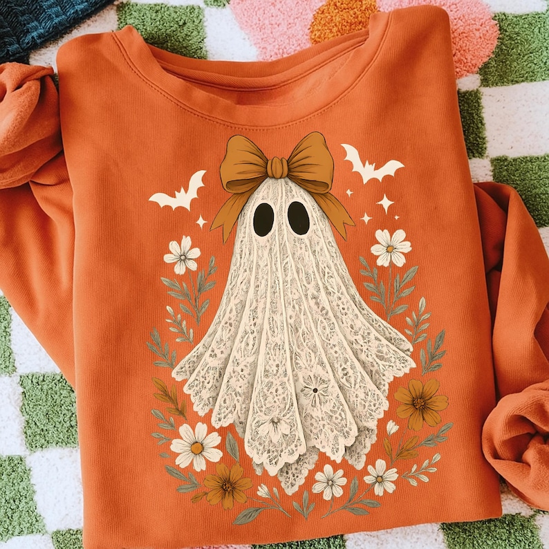 May include: Orange sweatshirt featuring a ghost design. The ghost is depicted with a white lace-like body, black oval eyes, and a brown bow. The design includes bats, flowers, and leaves, creating a Halloween theme.