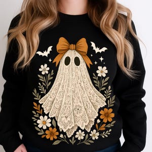 May include: Black sweatshirt featuring a Halloween ghost design. The ghost is made of white lace and has a brown bow. The design includes white bats, flowers, and stars. A festive and stylish apparel item for the spooky season.