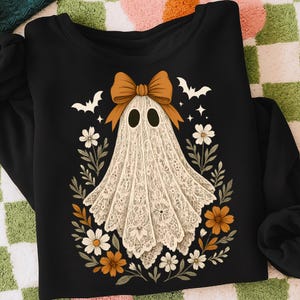 May include: Black sweatshirt featuring a ghost design. The ghost is white with a lace-like pattern, a brown bow, and surrounded by flowers and bats. A festive Halloween-themed apparel item.