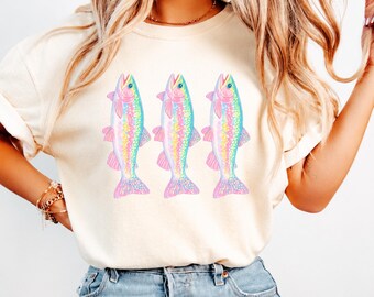 May include: A cream-colored short-sleeve t-shirt featuring a graphic print of three identical fish arranged side-by-side. Each fish is illustrated with a vibrant, multi-colored rainbow pattern, including shades of pink, yellow, blue, and purple.