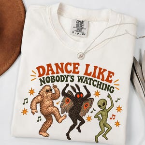 May include: A white t-shirt featuring a graphic of Bigfoot, a Mothman, and an alien dancing. The text reads DANCE LIKE NOBODY'S WATCHING in orange and dark green. The shirt has a Comfort Colors label at the neck and a small silver necklace.