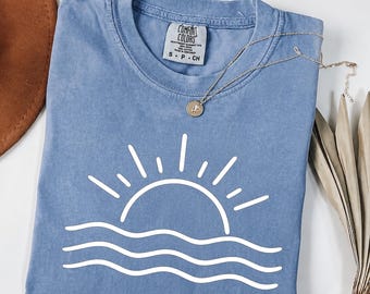 May include: A blue short-sleeved t-shirt featuring a white line art graphic of a sun rising over three wavy lines. The shirt has a Comfort Colors brand label at the inner neckline and is displayed with a silver chain necklace resting on the fabric.