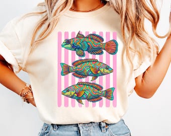 May include: A cream-colored short-sleeve t-shirt featuring a graphic print of three fish stacked vertically. Each fish displays a vibrant, psychedelic pattern of swirling neon colors. The fish are positioned over a background of vertical pink stripes.
