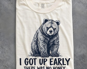May include: An off-white t-shirt featuring a dark blue graphic of a grumpy-looking bear sitting in grass. Below the bear, the text reads, "I GOT UP EARLY THERE WAS NO HONEY." The shirt has a crew neckline and a Comfort Colors brand label at the neck.