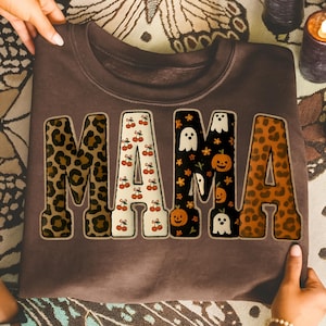 Pode incluir: Um moletom marrom com a palavra "MAMA" em letras grandes e decorativas. As letras têm padrões diferentes: estampa de leopardo, cerejas, fantasmas e abóboras, e estampa de leopardo. O moletom é segurado por mãos.