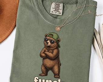 May include: An olive green t-shirt featuring a graphic of a brown bear wearing a green baseball cap and black sunglasses with arms crossed. The word "funcle" is printed in cream-colored letters below the bear. The shirt has a Comfort Colors label.