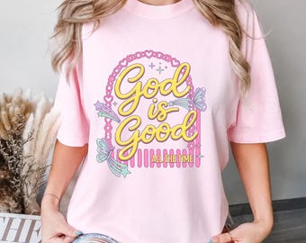 May include: A light pink short-sleeved t-shirt featuring a graphic print with the yellow text God is Good and All The Time. The design includes a pink arched frame, pastel-colored butterflies, and small star accents on the front of the shirt.