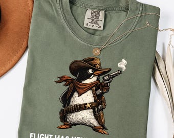 May include: An olive green t-shirt featuring a graphic of a penguin dressed as a cowboy, wearing a hat, scarf, and gun belt, holding a smoking revolver. The text FLIGHT WAS NEVER AN OPTION is printed in white below the graphic. The shirt has a Comfort Colors label.