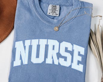 May include: A blue short-sleeved t-shirt with a vintage wash finish. The shirt features the word NURSE printed in a large, arched, white and light blue vertical striped font across the chest. A small gold-toned pendant necklace rests on the neckline.