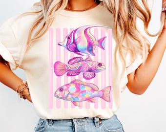 May include: A cream-colored t-shirt featuring a graphic print of three tropical fish in shades of pink, purple, and blue. The fish are arranged vertically against a background of soft pink vertical stripes, creating a colorful and detailed design.