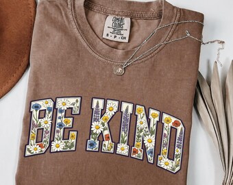 May include: A brown short-sleeved t-shirt with a crew neckline featuring the text BE KIND in large block letters filled with a colorful floral pattern of daisies, poppies, and cornflowers. A silver chain necklace with a small circular pendant rests on top.