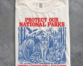 May include: An off-white t-shirt featuring a blue graphic of a bear, wolf, deer, raccoon, rabbit, and eagle in a forest setting. The red text reads PROTECT OUR NATIONAL PARKS and WHERE LEGENDS STILL ROAM FREE. The shirt has a Comfort Colors label.