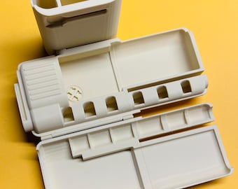 May include: A cream-colored plastic organizer case with a hinged lid and a separate upright cup component. The interior features multiple rectangular compartments and a row of five small circular slots for holding items securely.