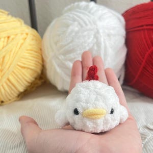 May include: A handmade, white crochet chicken with a red comb and yellow beak is held in a hand. Three balls of yarn in yellow, white, and red are in the background. The chicken is a small, plush toy.