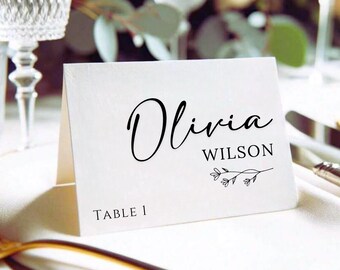 Pode incluir: Um cartão de lugar branco com o nome "Olivia Wilson" em elegante escrita preta. O cartão apresenta um raminho floral e o número da mesa "1" na parte inferior, ideal para um casamento ou evento.