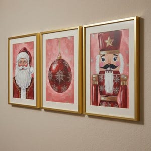 Pode incluir: Três impressões de arte emolduradas com tema natalino. As impressões apresentam o Papai Noel, uma bola vermelha com flocos de neve e um soldado quebra-nozes. Cada impressão está em um fundo rosa com detalhes prateados e emoldurada em dourado.