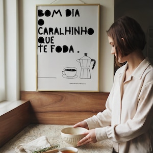 May include: A framed wall poster featuring black text that reads BOM DIA O CARALHINHO QUE TE FODA, accompanied by a simple black line drawing of a coffee cup and a moka pot. The poster is displayed in a thin gold frame against a plain white wall.
