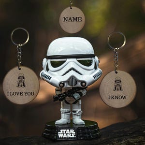 Peut inclure: Figurine de Stormtrooper Star Wars sur une base noire avec le logo Star Wars. Trois porte-clés en bois avec les textes "NAME", "I LOVE YOU" et "I KNOW" sont également présents. Les porte-clés ont une petite illustration de Dark Vador.