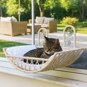 Pode incluir: Uma rede para gatos tecida em bege com uma almofada de tecido branco, fixada a uma janela de vidro com quatro ventosas transparentes. Um gato tigrado castanho descansa dentro da rede, que apresenta uma estrutura em treliça e dois braços arqueados.