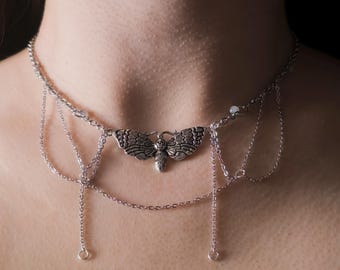 May include: Silver-toned choker necklace featuring a detailed moth pendant. The necklace has delicate chain drapes and small, round accents. The moth pendant is the focal point, with intricate wing and body details.