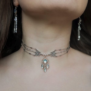Puede incluir: Collar gargantilla plateado con un diseño de tres hilos y un colgante central con tres gemas en forma de lágrima de color azul claro. También se ven pendientes colgantes a juego con gemas y detalles de cadena similares.