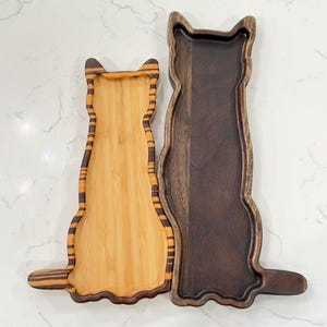 May include: Two wooden cat-shaped serving trays. One is made of light bamboo with dark brown stripes and trim. The other is a dark brown wood. Both trays have a recessed area for holding items. The trays are designed to resemble a cat.