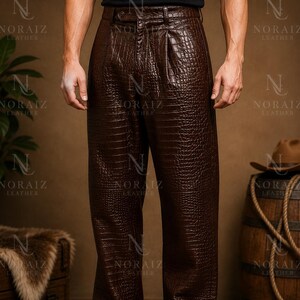 May include: Brown leather trousers with a crocodile texture. The trousers have a high waist and a pleated front. The model is wearing black shoes. The brand name "NORAIZ LEATHER" is visible in the background.