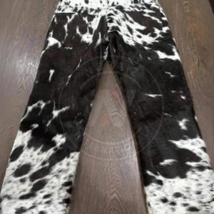 May include: Full-length pants featuring a black and white cow print pattern throughout. The garment includes a waistband with belt loops and two back pockets. The texture appears to have a short-haired finish, displaying a high-contrast spotted design across the entire surface of the trousers.