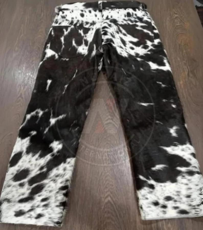 May include: Full-length pants featuring a black and white cow print pattern throughout. The garment includes a waistband with belt loops and two back pockets. The texture appears to have a short-haired finish, displaying a high-contrast spotted design across the entire surface of the trousers.