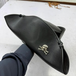 May include: A black tricorn hat featuring a smooth, matte finish and a prominent silver-toned skull and crossbones emblem centred on the front brim. The hat is constructed with folded edges secured by black cord detailing at the corners.