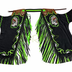 May include: Black leather chaps featuring neon green fringe and trim. The upper sections display brown embossed leather panels and embroidered patches of a skull wearing a feathered headdress. The garment includes adjustable side buckles.