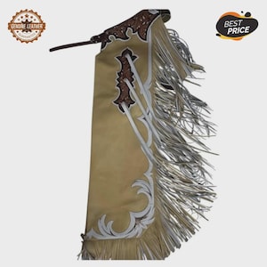 May include: Tan leather chaps with brown and white decorative elements and fringe. The chaps have a decorative design along the edges and a strap at the top. The words "Genuine Leather Guarantee" are in a circular logo.