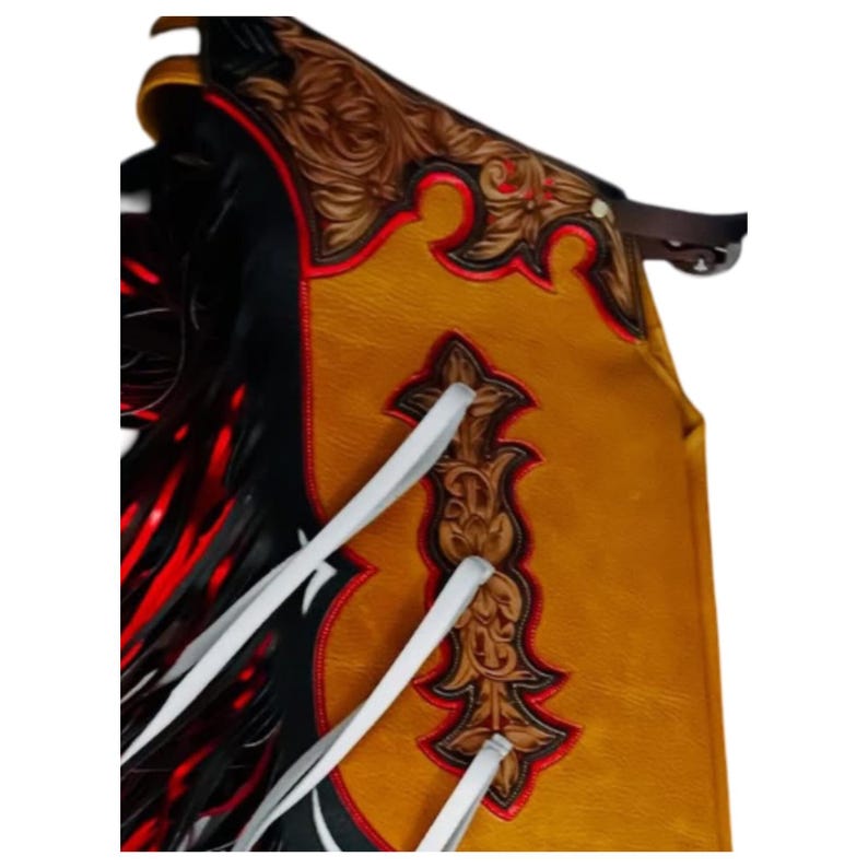 May include: Close-up of a western-style leather chaps. The chaps feature a tan and black color scheme with red accents. Intricate floral tooling and white ties add detail to the design. A brown leather strap is visible at the top.