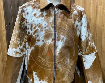 May include: A cowhide jacket with a brown and white spotted pattern. The jacket has a collar, a zipper closure, and brown leather accents on the shoulders and sleeves. The sleeves have fringe detailing.