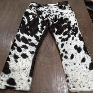 May include: Full-length pants featuring a black and white cow print pattern throughout. The garment includes a standard waistband with belt loops, a front button closure, and a classic five-pocket design with visible stitching along the seams.