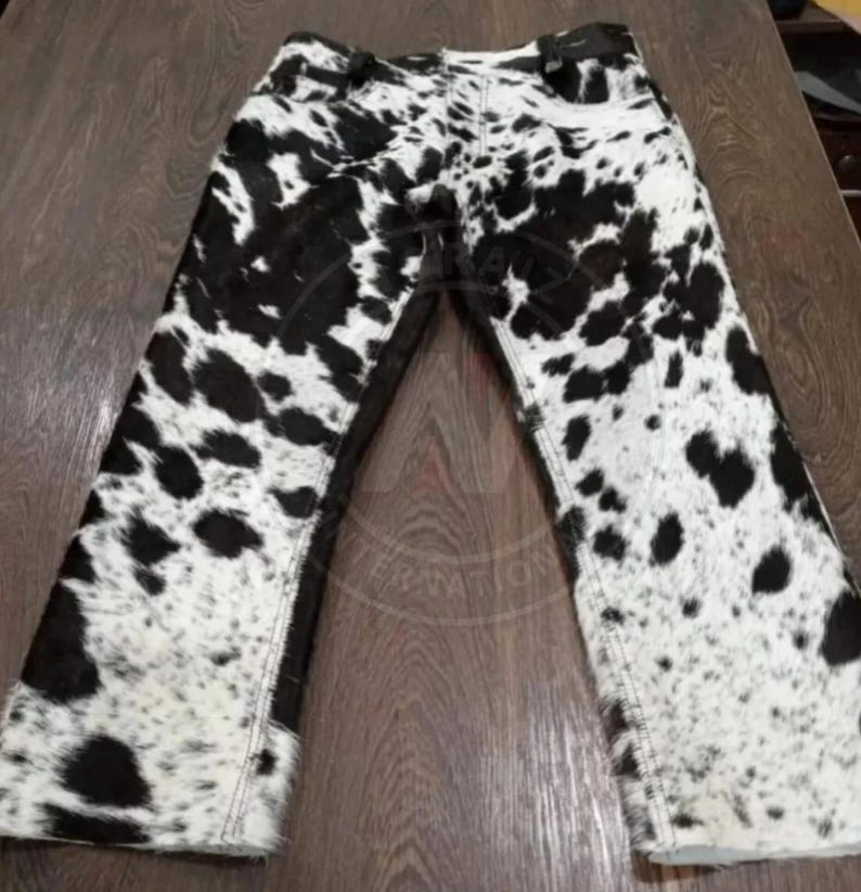 May include: Full-length pants featuring a black and white cow print pattern throughout. The garment includes a standard waistband with belt loops, a front button closure, and a classic five-pocket design with visible stitching along the seams.