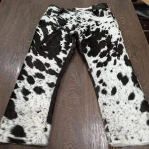 May include: A pair of pants featuring a black and white cow print pattern throughout. The garment has a standard waistband with belt loops, a front fly closure, and straight legs with visible stitching along the seams and hemline.