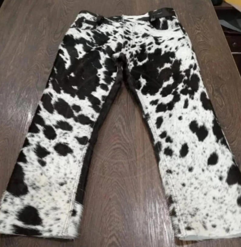 May include: A pair of pants featuring a black and white cow print pattern throughout. The garment has a standard waistband with belt loops, a front fly closure, and straight legs with visible stitching along the seams and hemline.