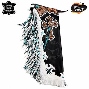 May include: Black leather chaps featuring white leather trim, long white and teal fringe, and a large embossed cross design on the upper thigh. The piece includes intricate floral tooling on the yoke and cross, with a belt strap at the top.