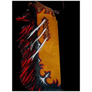 May include: Mustard yellow leather chaps with black and red fringe. The chaps have decorative white ties and a brown leather strap. The chaps have a decorative design in black and white.