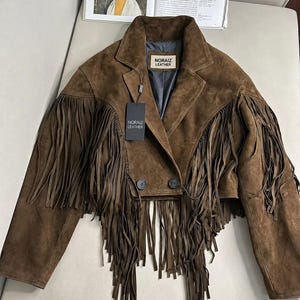 Puede incluir: Una chaqueta corta de ante marrón con largos flecos en el pecho y las mangas. La chaqueta tiene cuello de solapa, dos botones frontales y un forro interior gris oscuro. Incluye una etiqueta negra con el texto NORAIZ LEATHER.