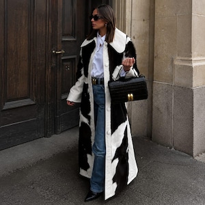 May include: A long coat featuring a black and white cow-print pattern with a large white collar, worn over a white button-down shirt and blue denim jeans. A black crocodile-embossed handbag with gold hardware is carried alongside the coat.