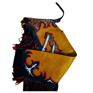 May include: Mustard yellow leather chaps with black and red accents. The chaps feature decorative floral and geometric patterns in brown and white. The chaps have a fringe detail and a black strap.