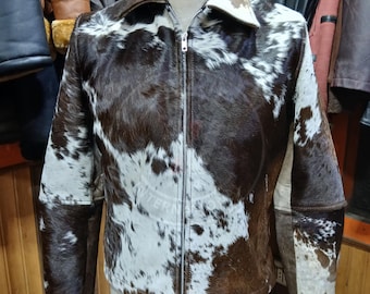 May include: A jacket made of cowhide featuring a brown and white spotted pattern. The garment has a pointed collar, long sleeves, and a full-length silver-toned metal zipper closure running down the center front. The texture is furry.