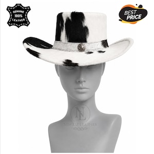 May include: A cow print cowboy hat featuring a white and black patterned hide texture. The hat includes a light-coloured band around the crown adorned with a central metallic star emblem. The hat is displayed on a grey mannequin head.
