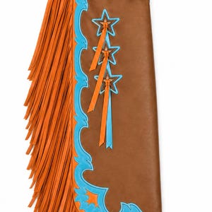 May include: A brown leather chap featuring long orange fringe, bright blue scalloped trim, and three star-shaped cutouts with dangling orange and blue ribbons. The upper section displays intricate floral tooling and a buckle closure strap.