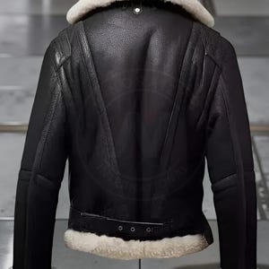 May include: Black leather jacket with a cream-colored shearling collar and trim. The jacket features a belted waist, a zipper closure, and a classic aviator style. The back of the jacket has a distinctive V-shaped design.