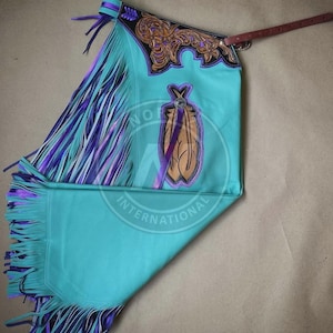 May include: Turquoise leather chaps with purple and turquoise fringe. The chaps feature a decorative floral design in brown and black, and a feather design. A brown leather belt is attached to the chaps.
