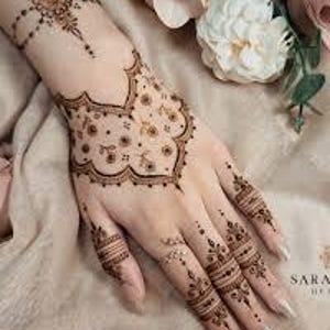 May include: Intricate henna designs adorn a hand, featuring floral and geometric patterns in brown. The detailed artwork extends from the wrist to the fingertips, with delicate dots and lines creating an ornamental effect. The background includes soft, neutral tones and floral accents.