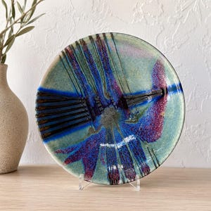 May include: A round ceramic plate featuring a variegated glaze in shades of sage green, deep cobalt blue, and magenta. The surface displays a series of dark, radiating linear patterns and textured splatters that converge towards the centre.